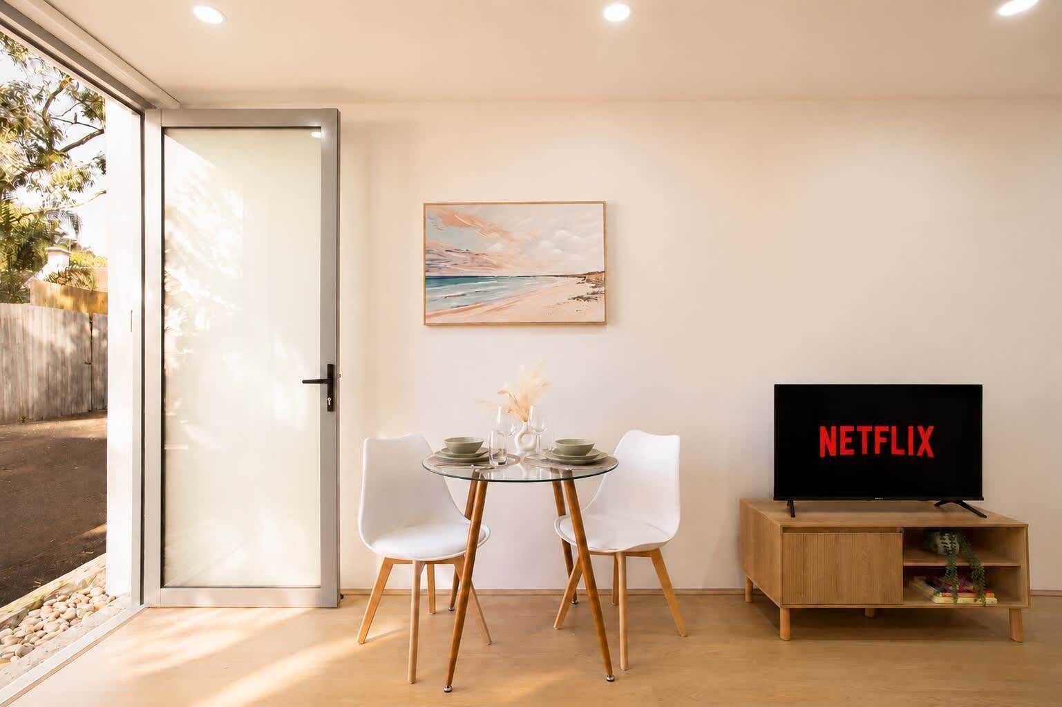 A modern living room with a glass dining table, white chairs, a wooden cabinet, a flat-screen TV displaying 'NETFLIX', and a beach-themed painting on the wall.