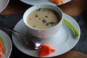 A bowl of soup on a white plate with a spoon, accompanied by a carrot and a green vegetable, possibly a cucumber, on a dark surface.