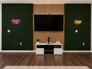 A modern living room with a large flat-screen TV mounted on a wooden panel wall, flanked by green foliage walls with neon signs. - Luxury King Suite • Beauty Room • Infinity Game Table • Near Hospital & ATL (Stockbridge)