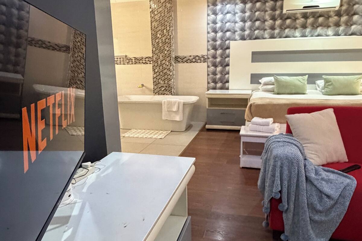 A hotel room with a bed, a red sofa, a TV, a bathroom with a bathtub, and a wall with a patterned design.