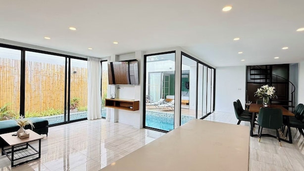 A modern interior with a large open space, a dining area, a TV mounted on a wall, and a view of a swimming pool.