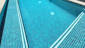 Ein Schwimmbecken mit Fliesenbord, eine überdachte Terrasse mit Stühlen und ein großes Fenster mit Vorhängen.