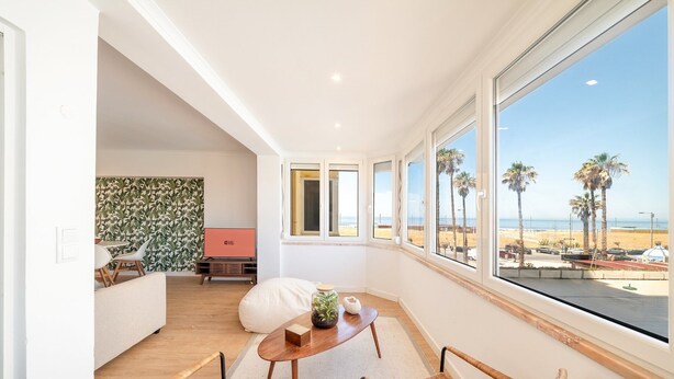 A modern living room with a large window, a sofa, a coffee table, and wicker chairs. - GuestReady - Stay at Costa da Caparica (Costa de Caparica)