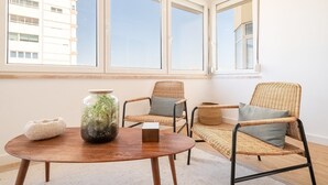 A room with a round wooden table, two wicker chairs with cushions, a glass vase with plants, and a view of a building through large windows. - GuestReady - Stay at Costa da Caparica (Costa de Caparica)
