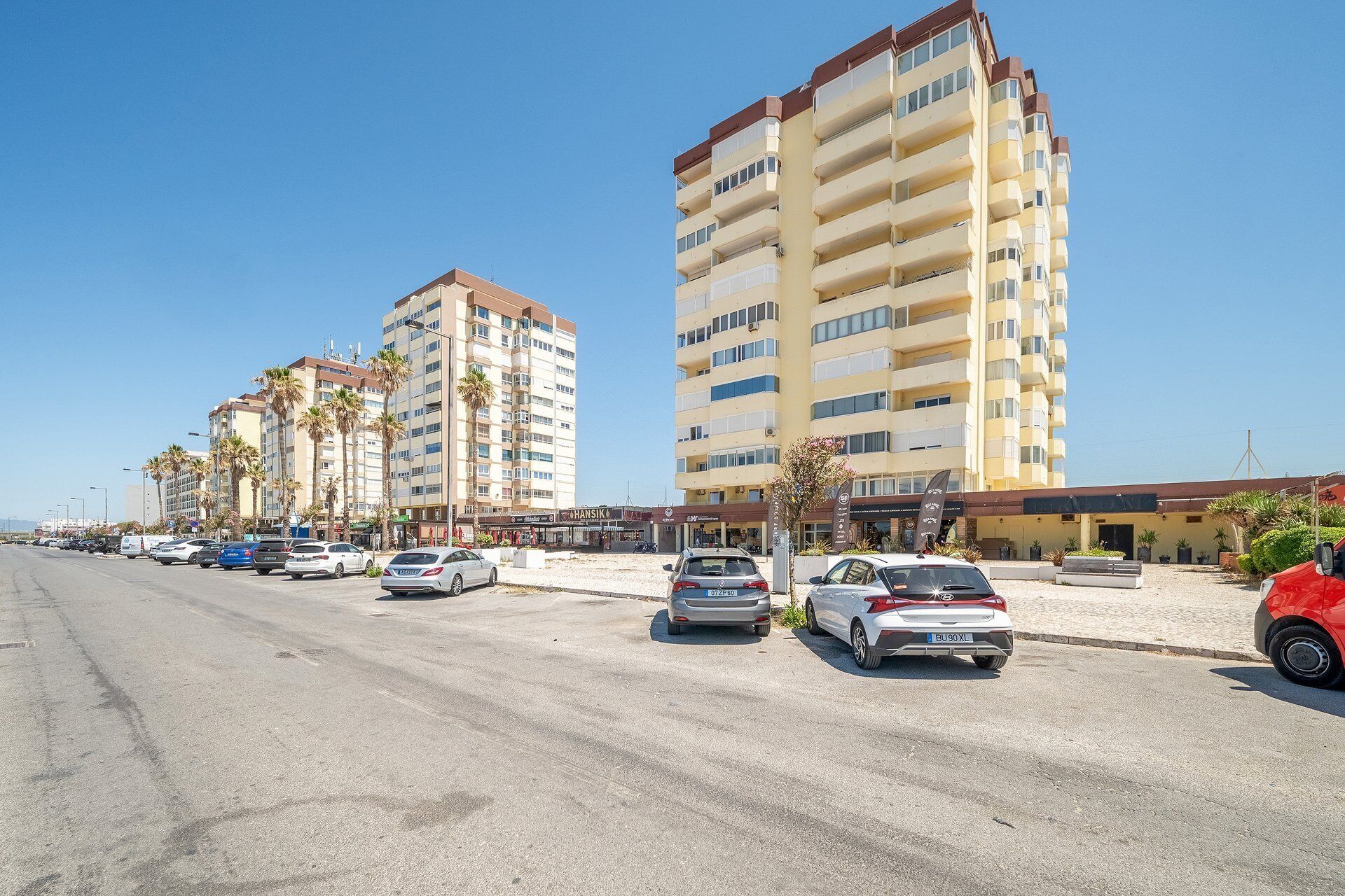 Un hôtel situé sur la côte, avec plusieurs étages, un parking où sont garées plusieurs voitures et des palmiers.