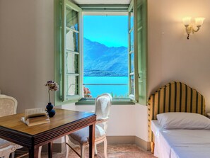 A room with a bed, a table with a book, a chair, and a window with a view of mountains and water.