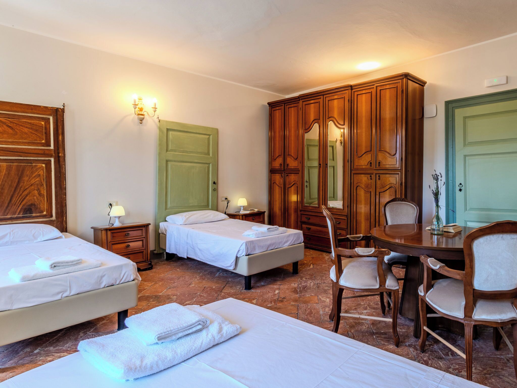 A hotel room with two beds, a wooden wardrobe, a dining table with chairs, and a green door.