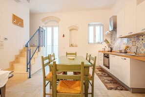 A dining area with a wooden table and chairs, a kitchen with white cabinets and a tiled backsplash, and a staircase with blue railings. - A wonderful home in the historic district (Castellana Grotte)