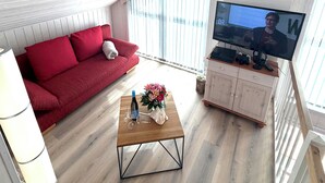 Ein Wohnzimmer mit einem roten Sofa, einem Holztisch mit einer Vase und einer Weinflasche sowie einem Fernseher auf einem Schrank.