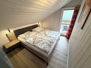 Ein Schlafzimmer mit holzverklebter Schräge, einem Bett mit floralem Bettzeug, einem kleinen Nachttisch und einem Fenster mit Vorhängen.