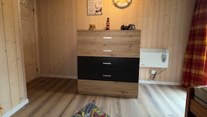 Ein Zimmer mit Holzwänden, einer hölzernen Kommode, einem an der Wand befestigten Rettungsring, einem kleinen Regal mit einer Flasche und einem Radio sowie einem weißen Heizgerät an der Wand.