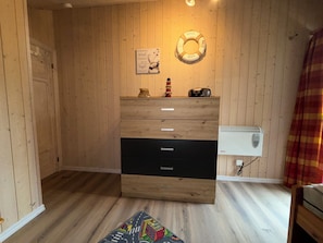Ein Zimmer mit Holzwänden, einer hölzernen Kommode, einem an der Wand befestigten Rettungsring, einem kleinen Regal mit einer Flasche und einem Radio sowie einem weißen Heizgerät an der Wand.