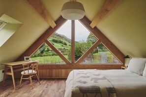 Ein Schlafzimmer mit großem Fenster, Holzbalken, einem Bett, einem Sessel und einem Schreibtisch.