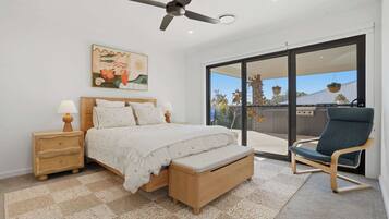 Une chambre à coucher comprenant un lit, des tables de chevet, un ventilateur de plafond, un tableau et un balcon avec une chaise.