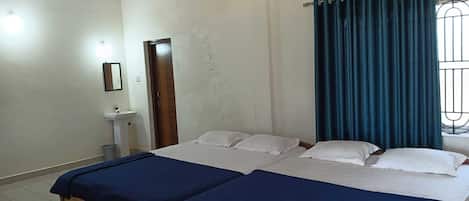 Een kamer met twee bedden, een plafondventilator, een airconditioning, een raam met gordijnen en een klein wastafeltje.
