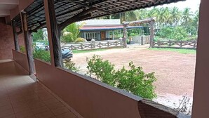 A covered corridor with a view of a fenced outdoor area, a building with a red roof, and a palm tree. - Stone Wood Stay (Bhatkal)