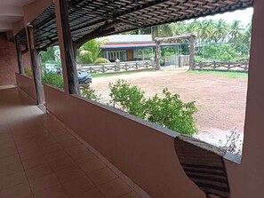 A covered corridor with a view of a fenced outdoor area, a building with a red roof, and a palm tree.