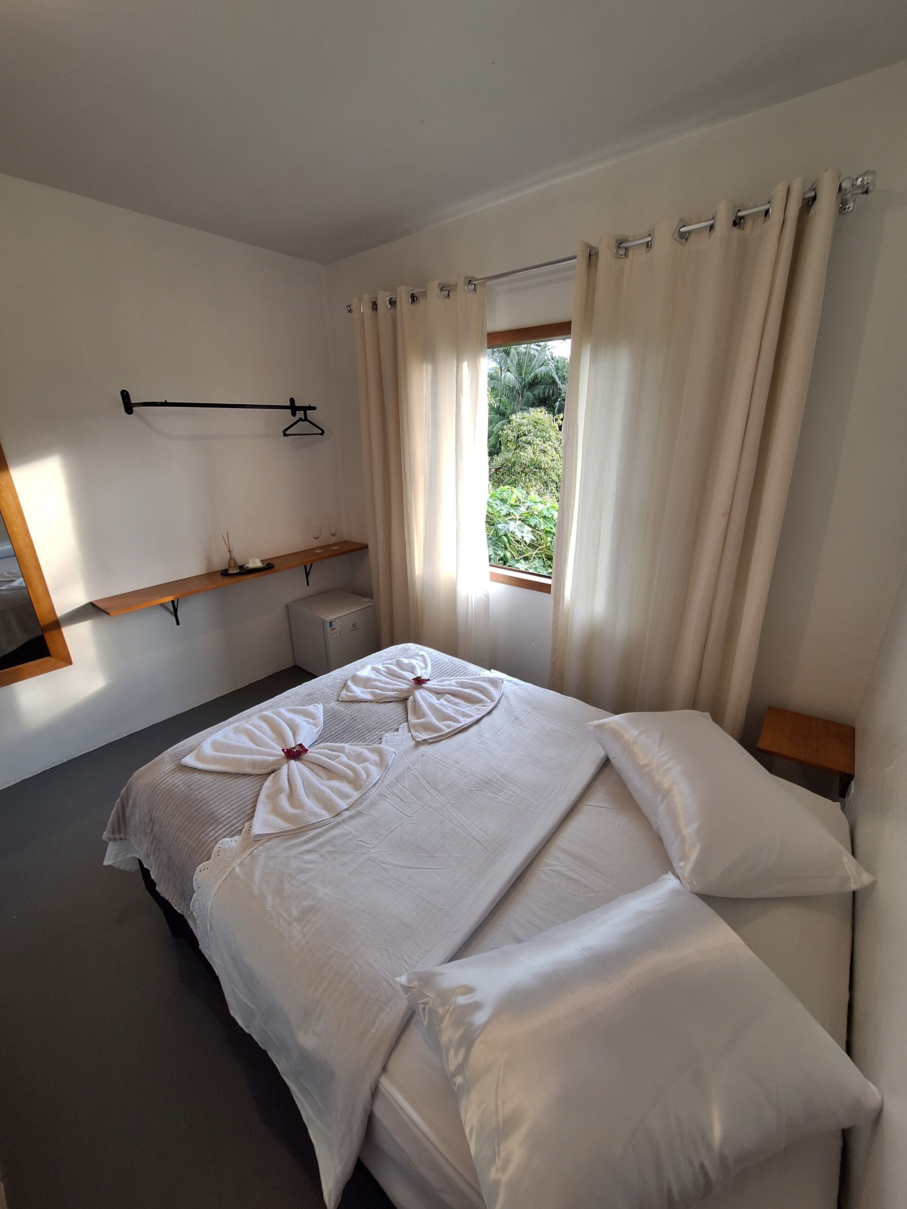 Una habitación de hotel con cama, mesita de noche, perchero y ventana con cortinas.