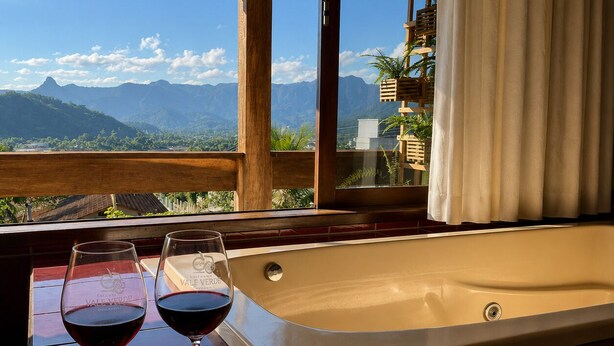 A room with a bathtub, a dining table with wine glasses and a fruit platter, and a view of mountains through large windows. - SOLAR DO VALE VERDE (Corupá)
