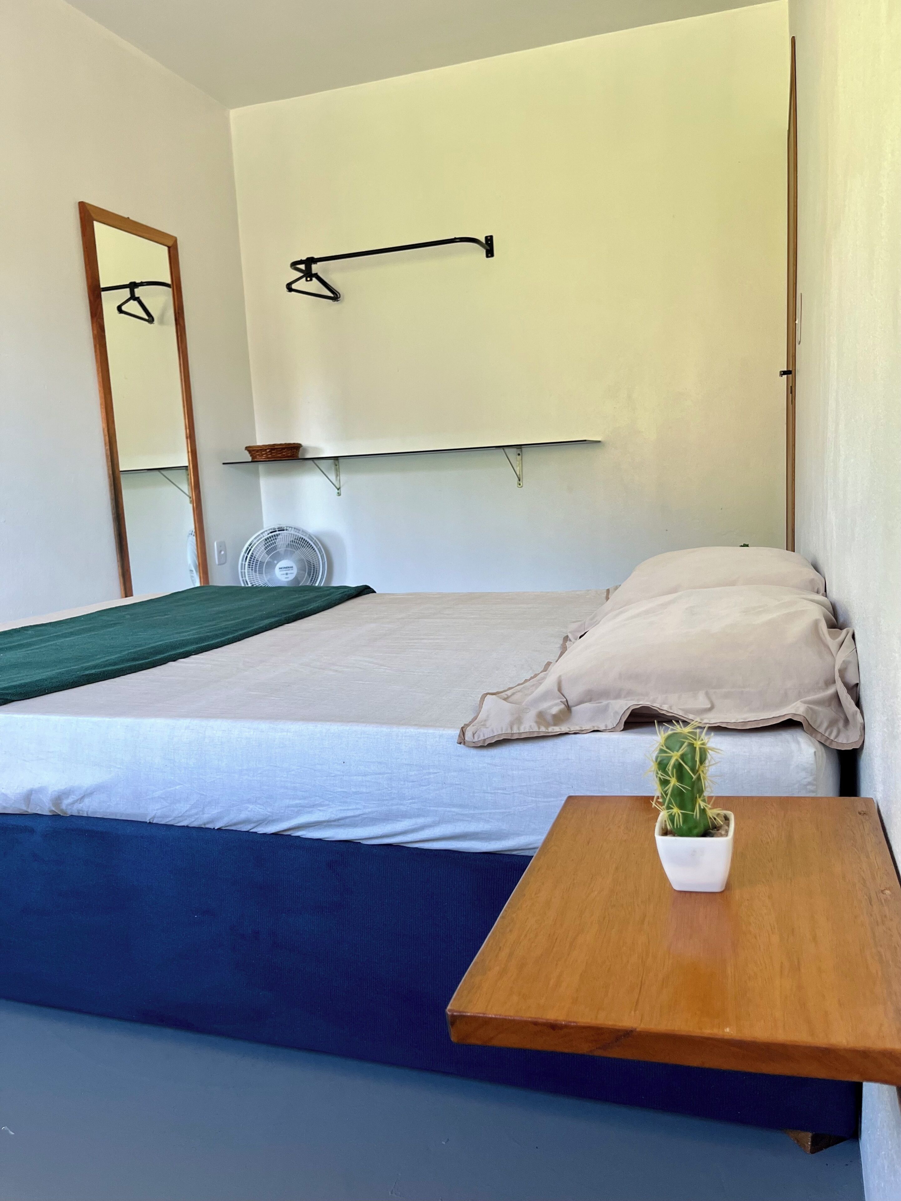 Ein Einzelbett mit weißer Matratze, ein Nachttisch aus Holz mit einem Topf mit Kakteen, ein Wandventilator und ein Spiegel.