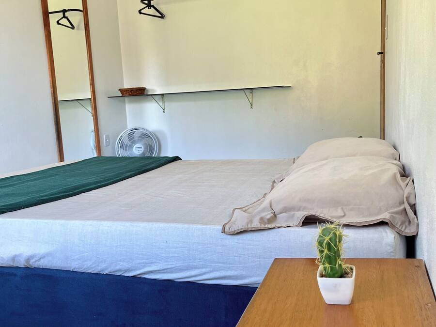 Ein Einzelbett mit weißer Matratze, ein Nachttisch aus Holz mit einem Topf mit Kakteen, ein Wandventilator und ein Spiegel.