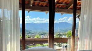 A room with a bed, white curtains, and a view of mountains and a town. - SOLAR DO VALE VERDE (Corupá)