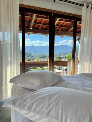 Une chambre avec un lit, des rideaux blancs et une vue sur les montagnes et une ville.