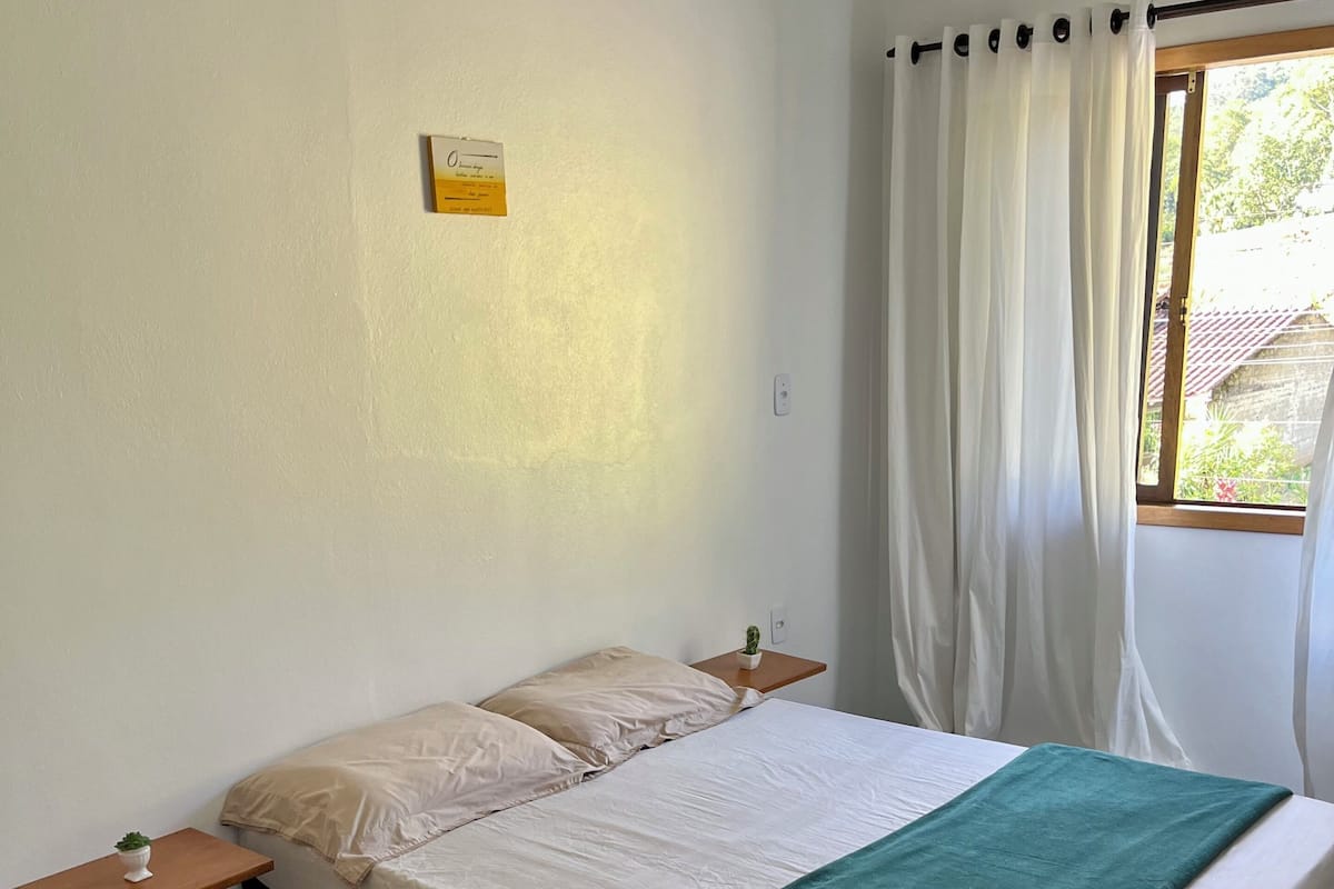 Una cama individual con una manta verde, una mesita de noche con una planta y una ventana con cortinas blancas.
