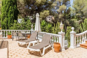 Ein sonnendurchfluteter Außenbereich mit Liegestühlen und einem Tisch, umgeben von einer weißen Balustrade und Blumentöpfen.
