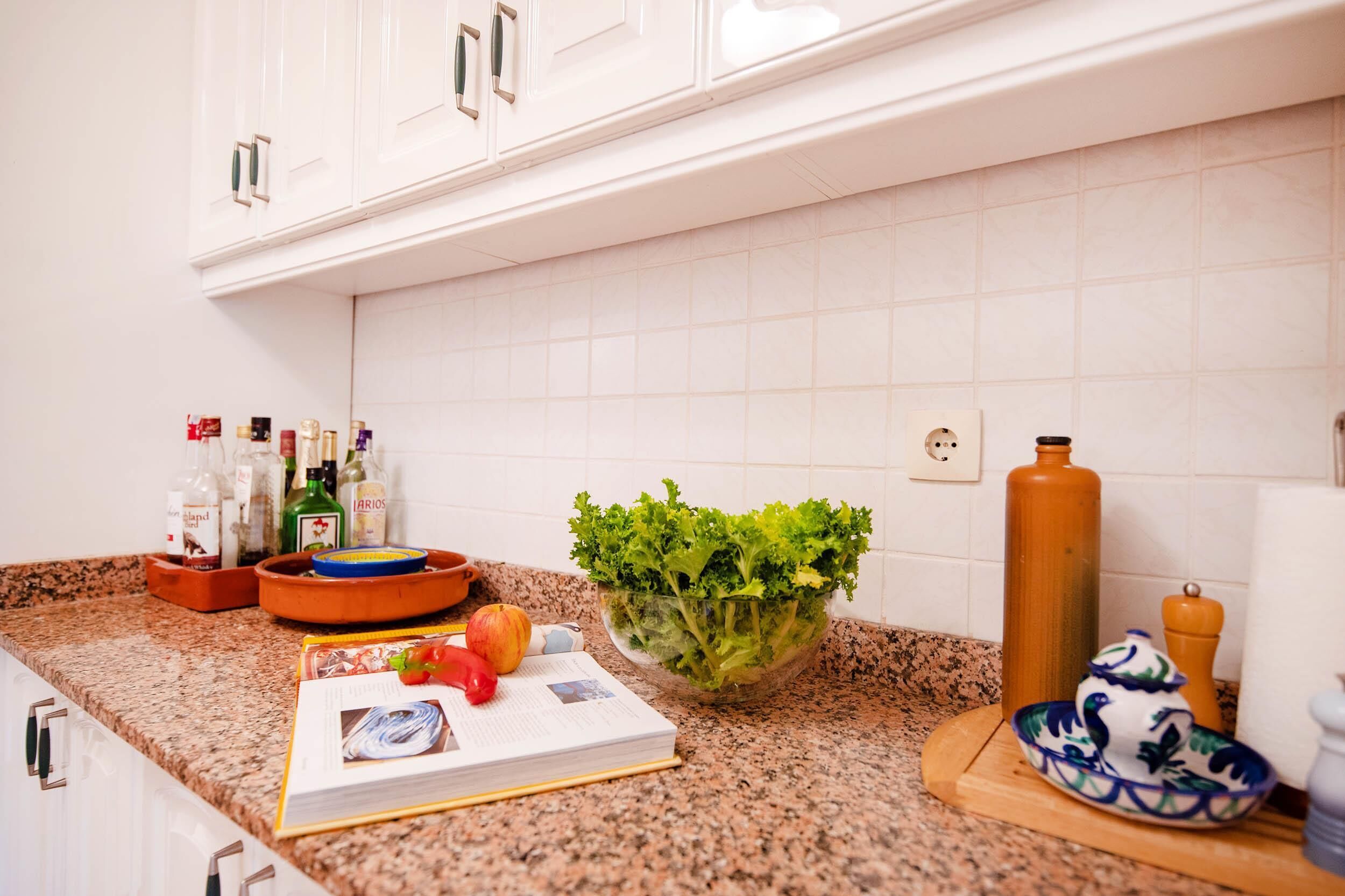 A kitchen with white cabinets, a granite countertop, a bowl of green salad, a book, and various kitchen items.