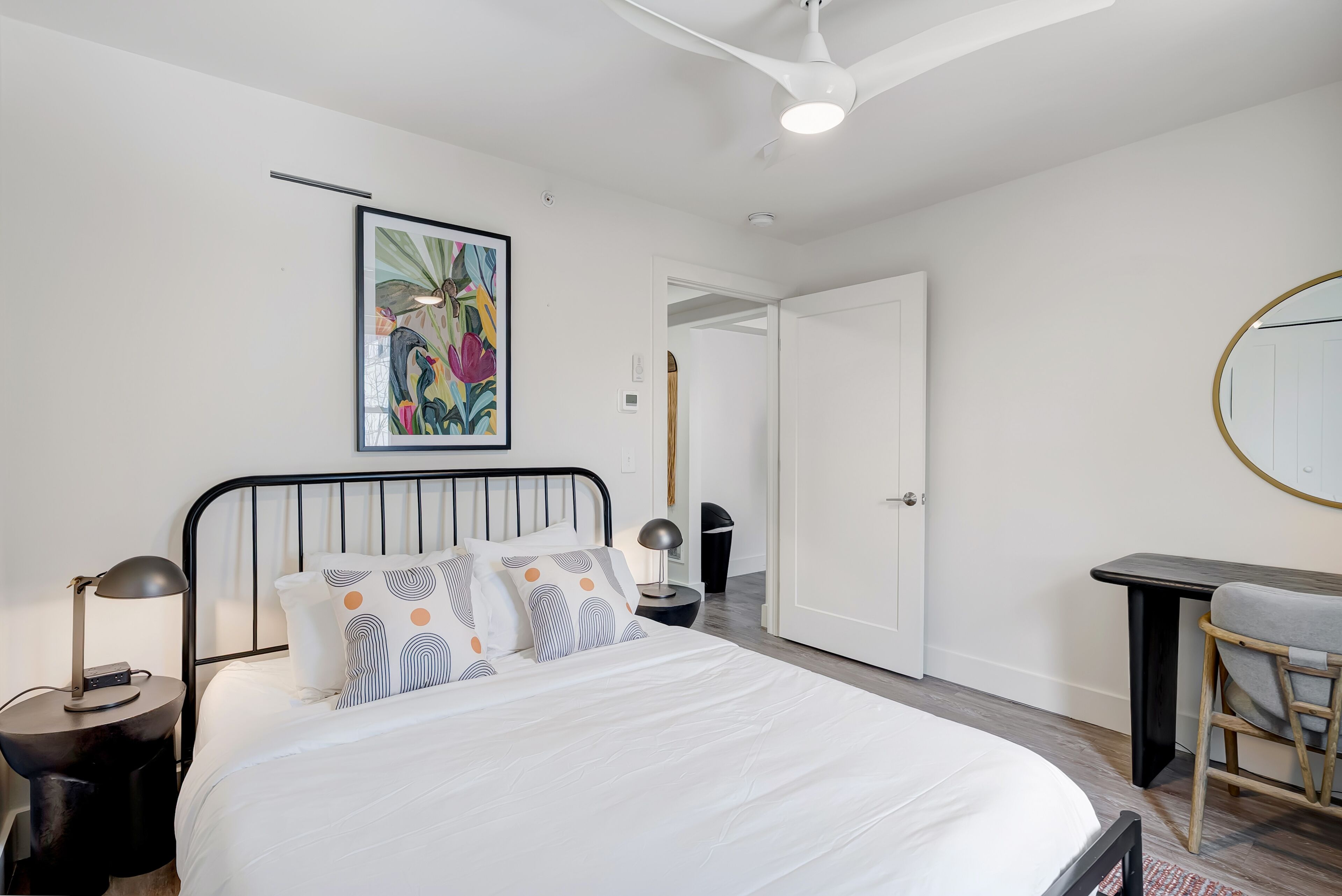 A bedroom with a bed, bedside tables, a desk, a chair, a ceiling fan, and a door.