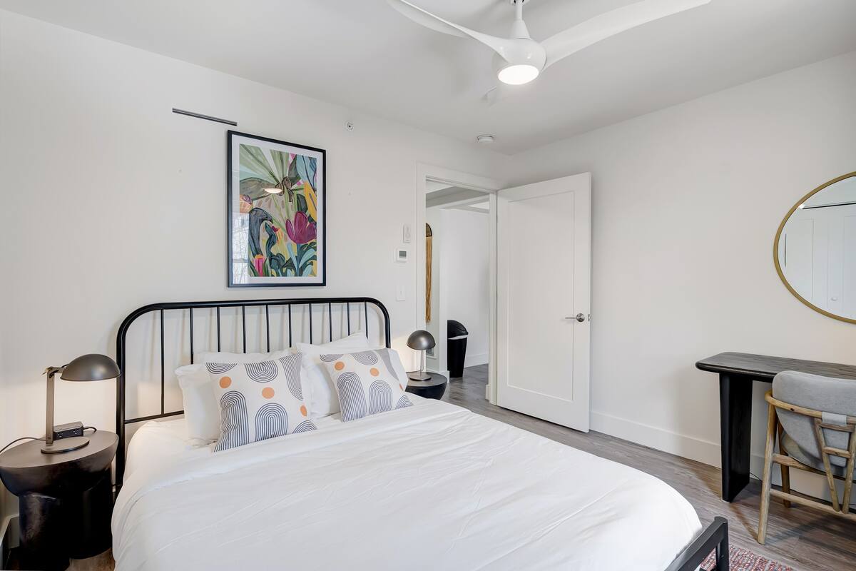 A bedroom with a bed, bedside tables, a desk, a chair, a ceiling fan, and a door.