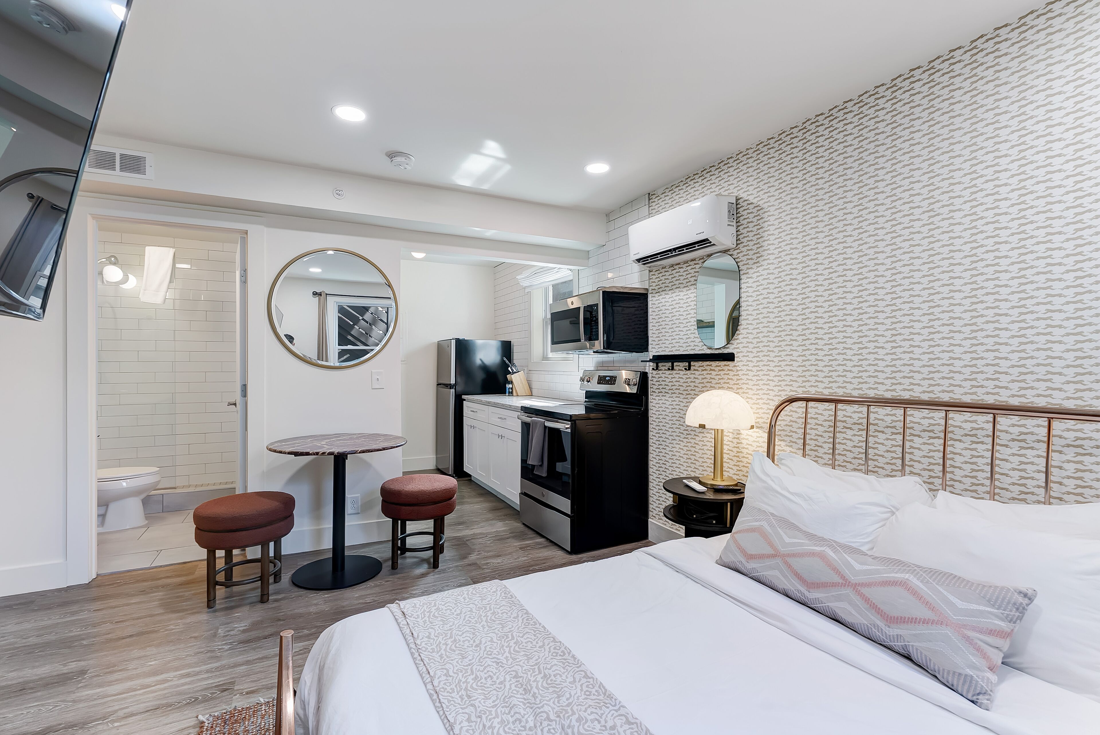 Une chambre d’hôtel moderne équipée d’un lit, d’une petite kitchenette, d’un miroir rond et d’une salle de bain avec des toilettes et un lavabo.