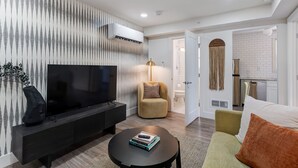 Una sala de estar moderna con un televisor de pantalla plana, un sofá, una mesa de centro y una cocina visible a través de una puerta abierta.