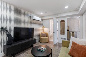 Una sala de estar moderna con un televisor de pantalla plana, un sofá, una mesa de centro y una cocina visible a través de una puerta abierta.