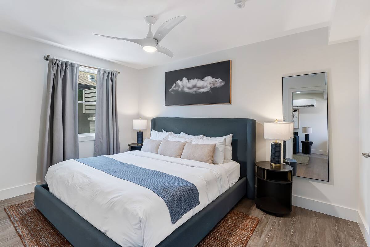 A modern bedroom with a bed, a ceiling fan, a window with curtains, a bedside table with a lamp, and a wall-mounted mirror.