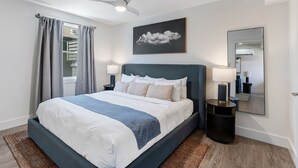 Un dormitorio moderno con cama, ventilador de techo, ventana con cortinas, mesita de noche con lámpara y espejo montado en la pared.