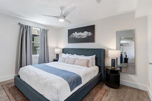 Un dormitorio moderno con cama, ventilador de techo, ventana con cortinas, mesita de noche con lámpara y espejo montado en la pared.