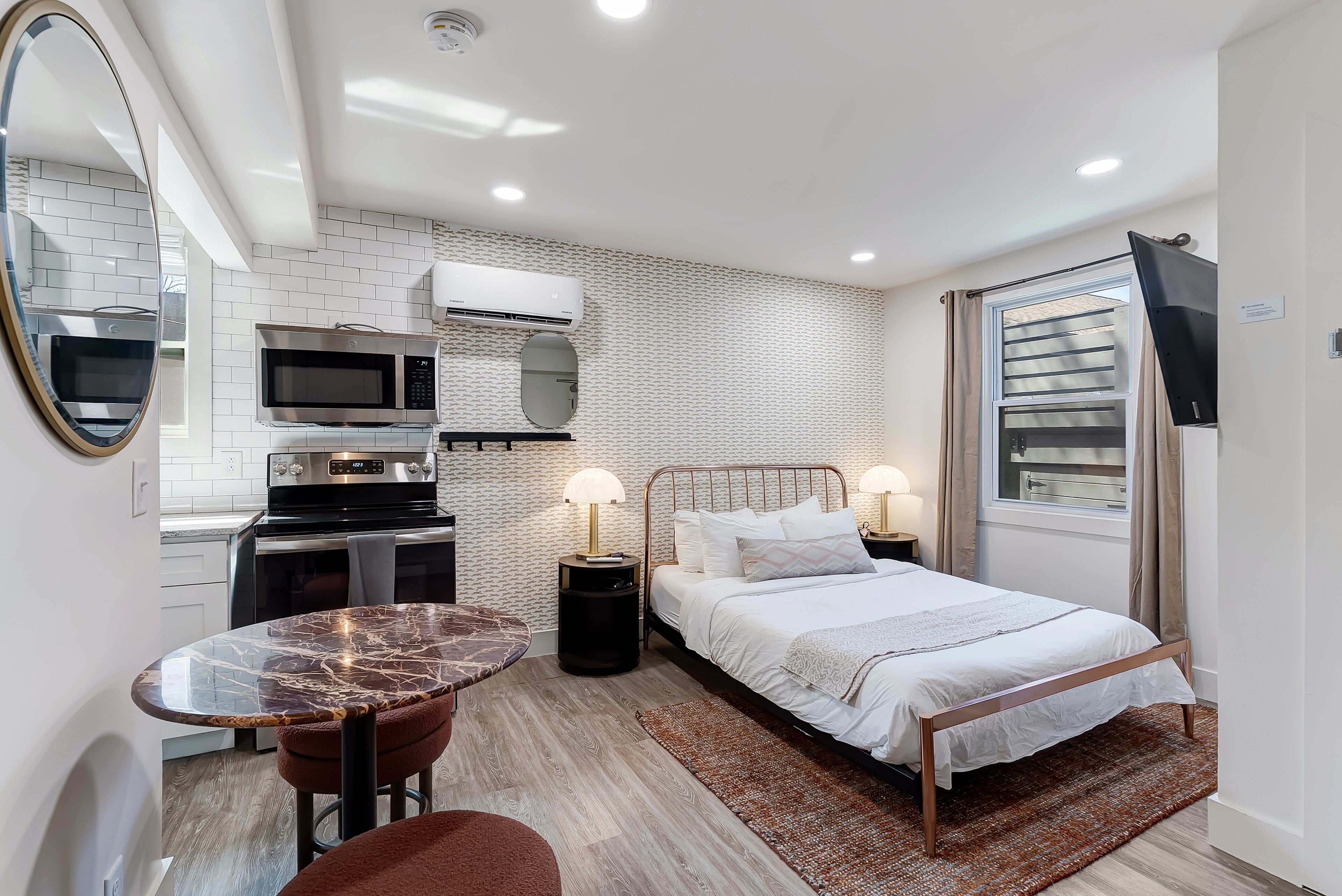 A compact room with a bed, bedside lamps, a small table, a mirror, a microwave, and an air conditioning unit.