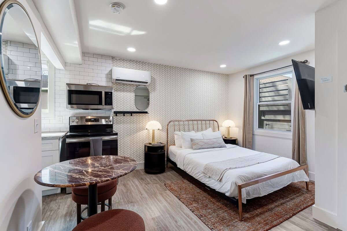 A compact room with a bed, bedside lamps, a small table, a mirror, a microwave, and an air conditioning unit.