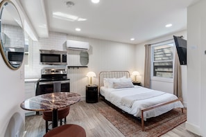 A compact room with a bed, bedside lamps, a small table, a mirror, a microwave, and an air conditioning unit. - State Street (Nashville)