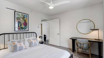 Un dormitorio con cama, escritorio, silla, ventilador de techo, espejo redondo y un cuadro en la pared.
