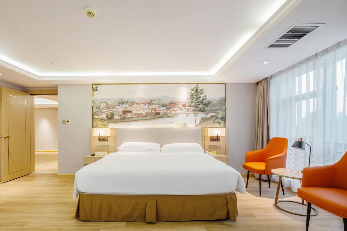 Quarto de hotel com uma cama grande, duas mesas de cabeceira, uma escrivaninha com cadeira, uma janela com cortinas e um quadro de paisagem urbana na parede.
