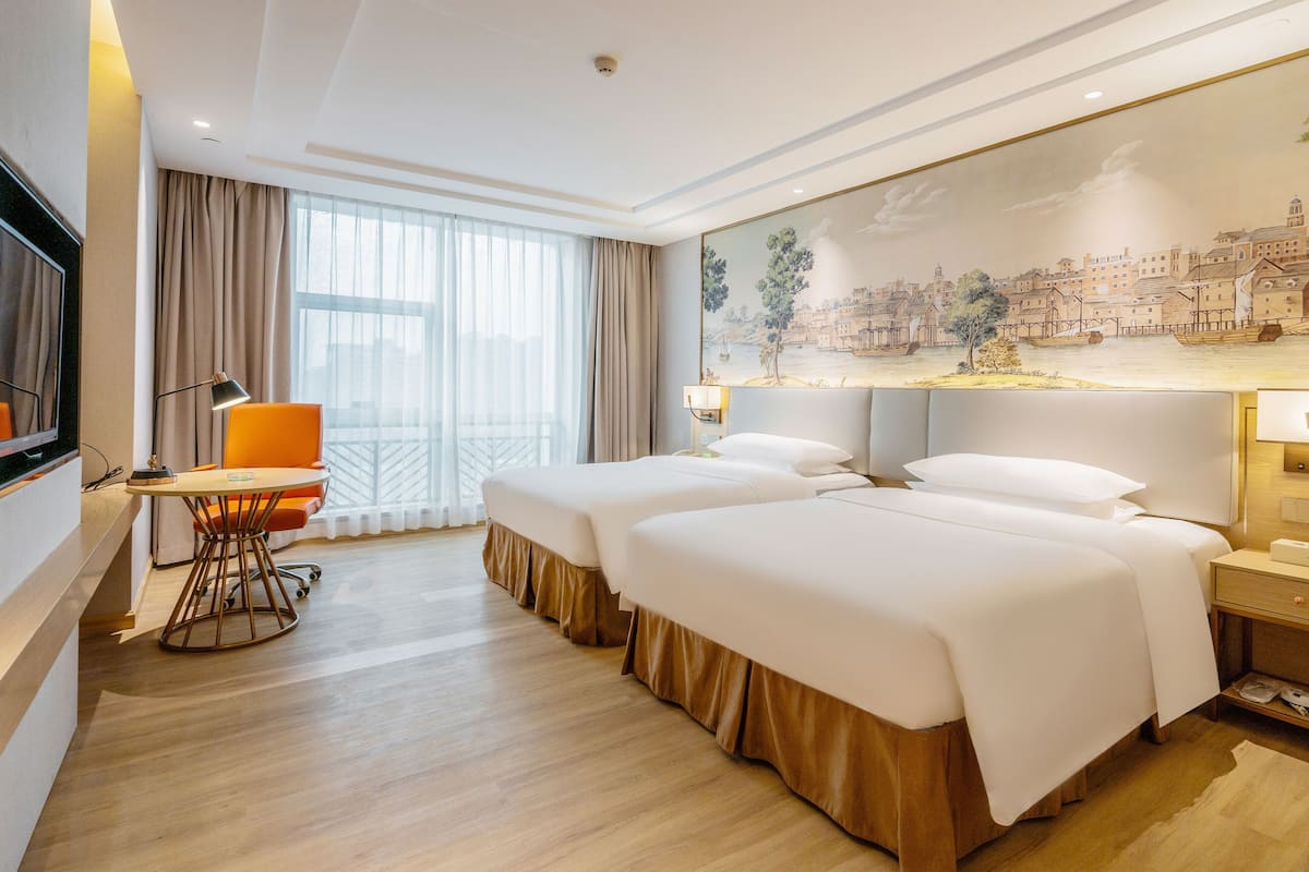 Quarto de hotel com duas camas, uma escrivaninha, uma cadeira, uma televisão e uma janela grande com cortinas.