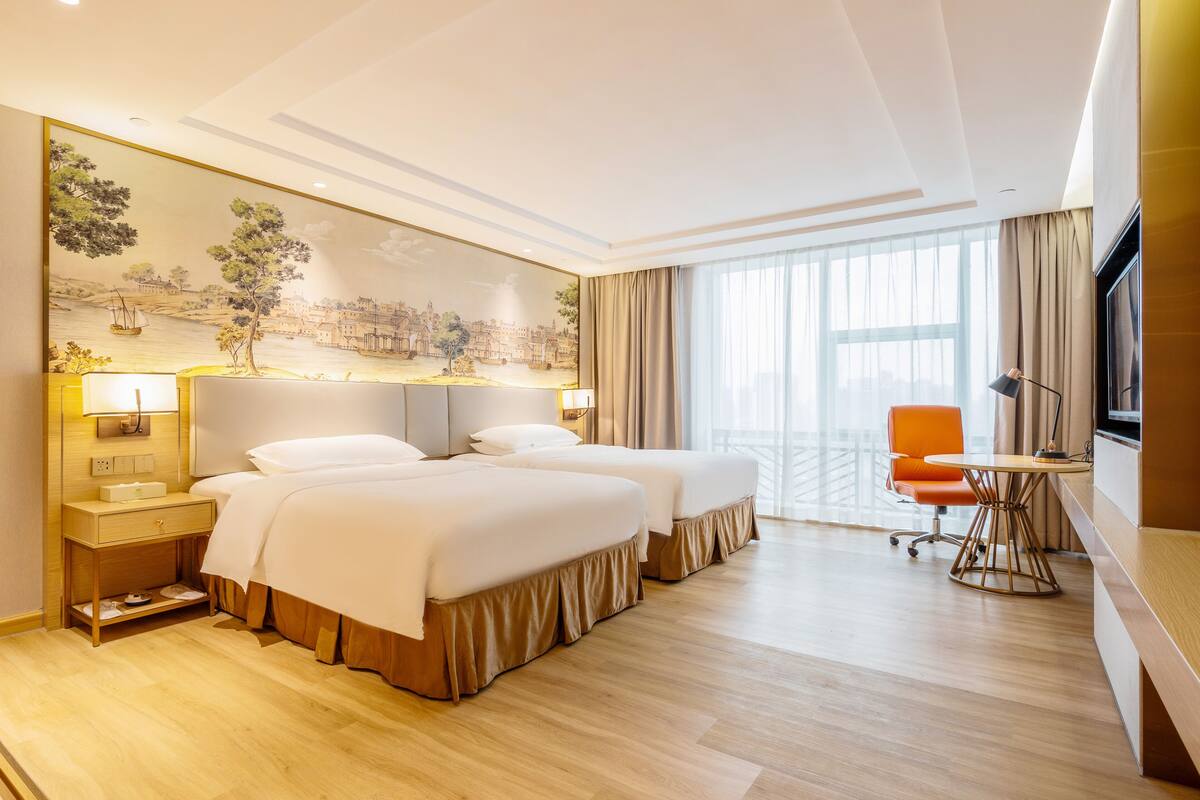 Quarto de hotel com duas camas, uma escrivaninha, uma cadeira, uma televisão e uma janela grande.