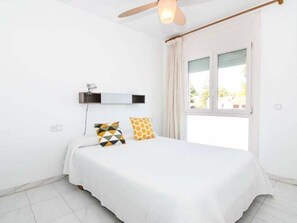 A bedroom with a bed, a ceiling fan, a window with curtains, and a wall-mounted shelf. - Apartament Puig - Comarruga (Comarruga)