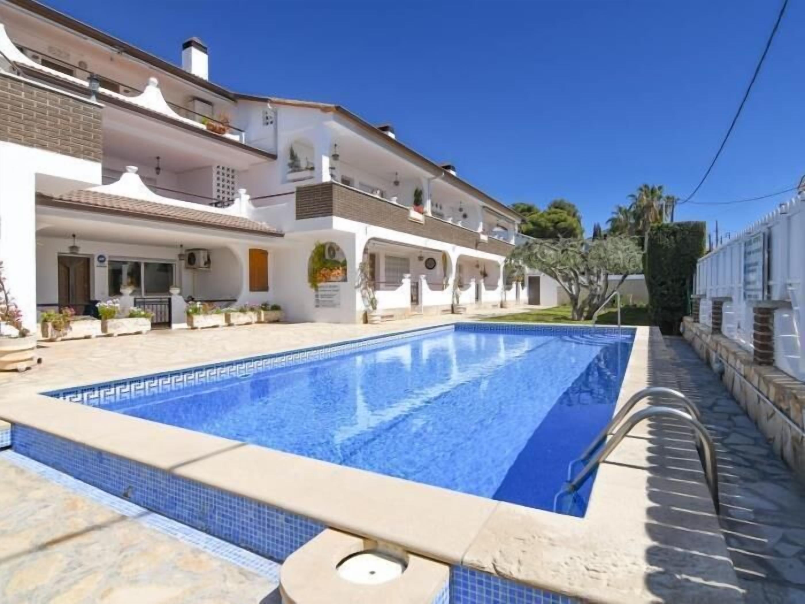 Una piscina amb un videt de rajola, un edifici blanc amb balcons i un cel blau clar.