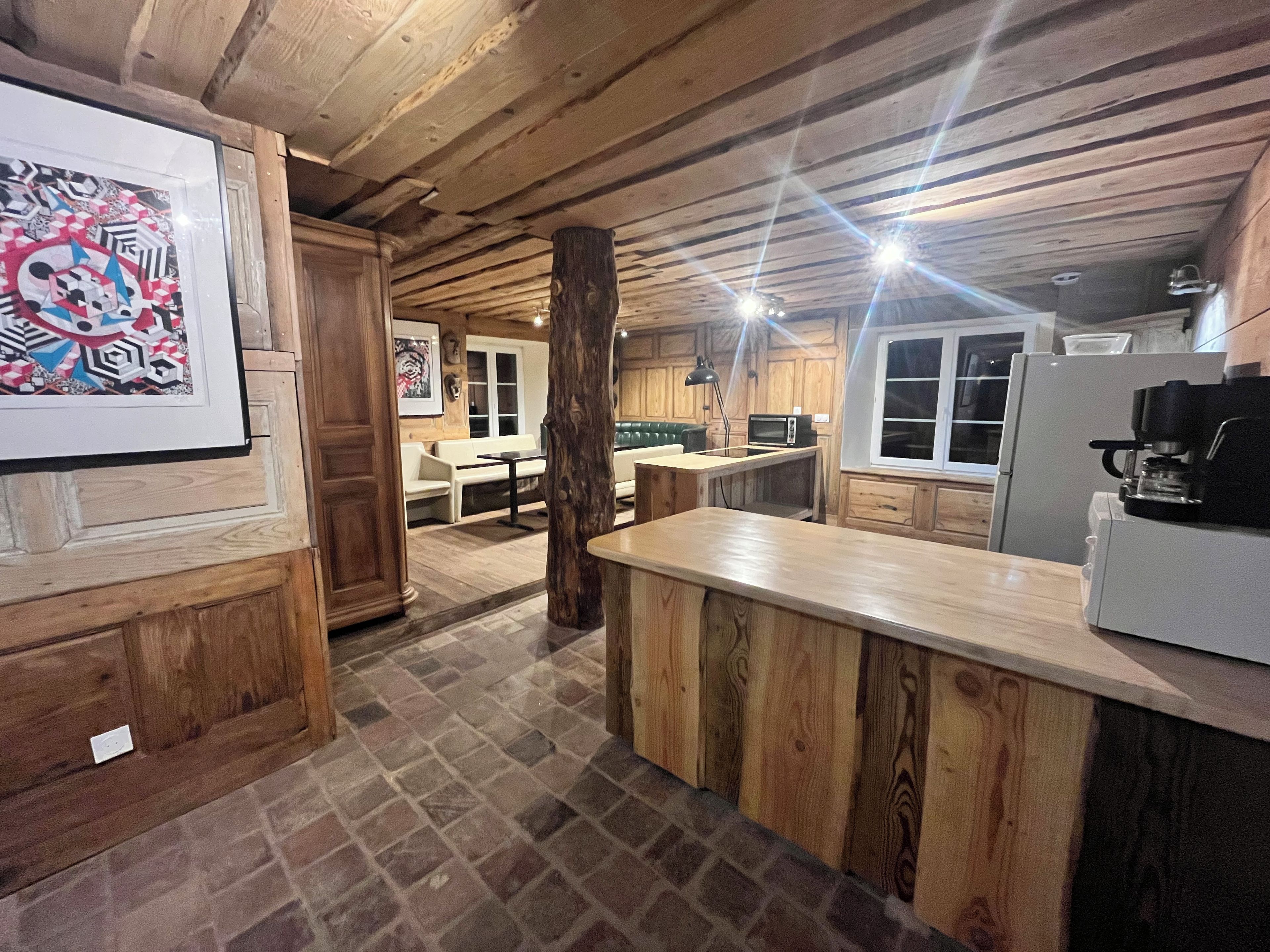 Uma cozinha rústica com vigas de madeira, piso de pedra, sala de jantar com mesa e cadeiras brancas, e uma geladeira.