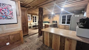 Uma cozinha rústica com vigas de madeira, piso de pedra, sala de jantar com mesa e cadeiras brancas, e uma geladeira.