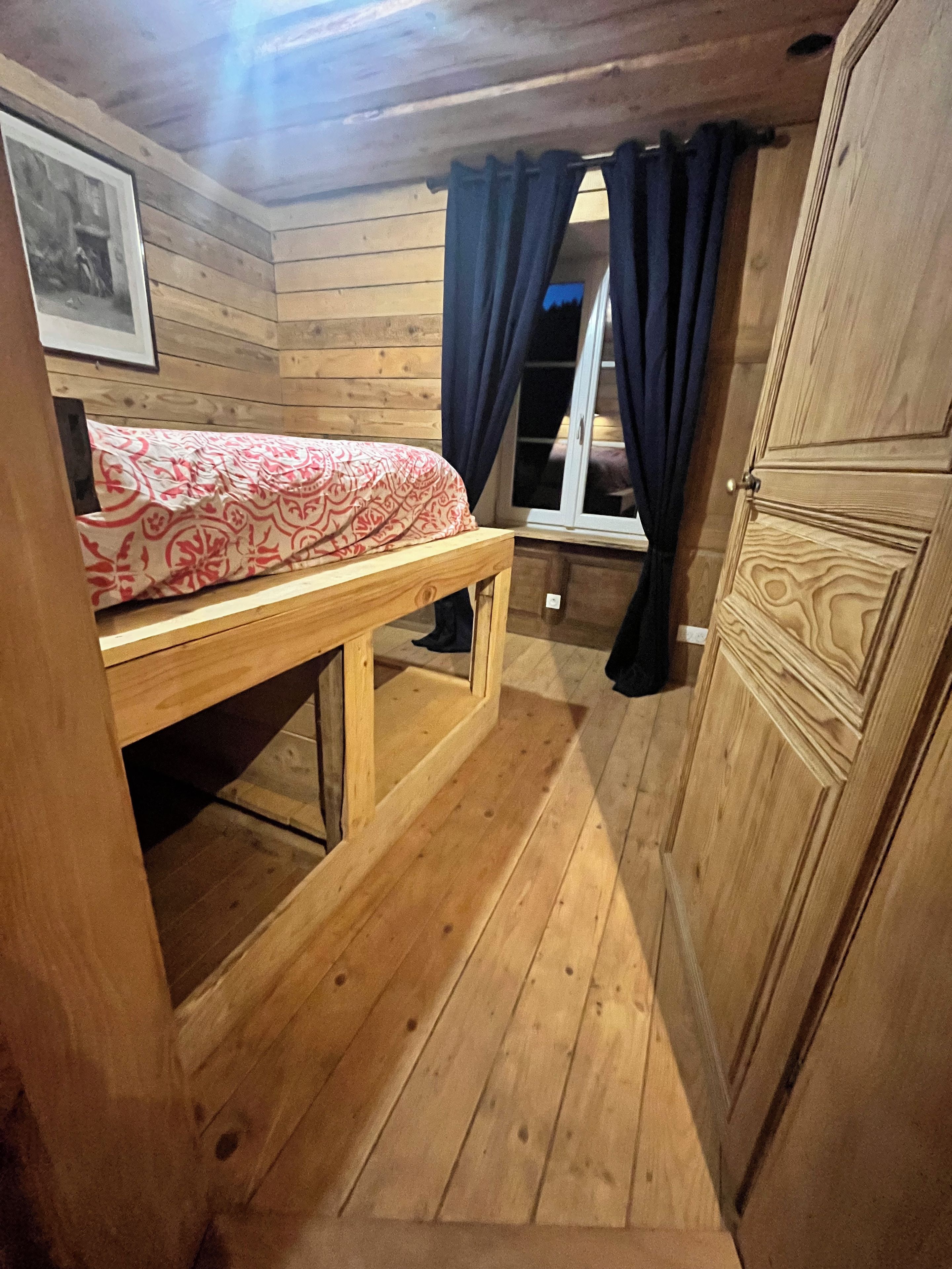 Ein Zimmer aus Holz mit einem Bett, einem Fenster mit Vorhängen und einer Tür.
